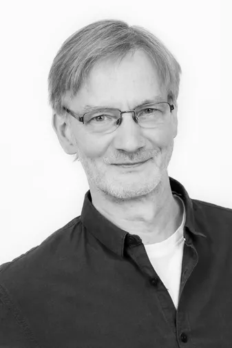 Black and white photo of an older man with glasses, short hair, and a beard, wearing an open-collar dark shirt and a light undershirt.