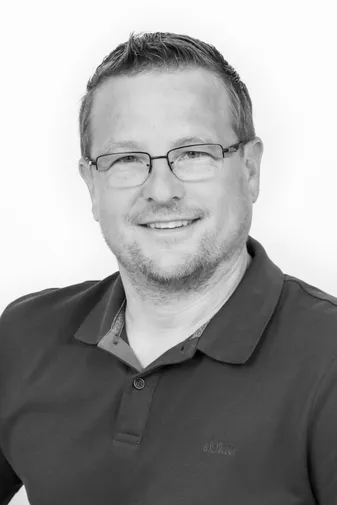 A person with short hair and glasses smiles while wearing a dark polo shirt. The photo is in black and white.