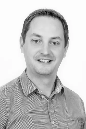 A man with short hair smiles at the camera, wearing a checkered button-up shirt against a white background. Black and white photo.