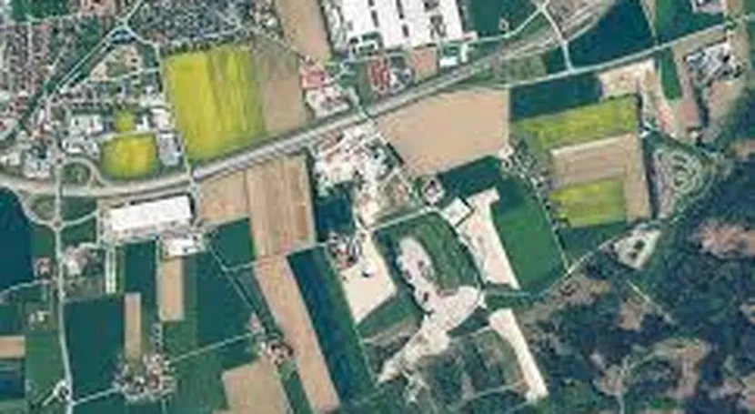 Aerial view of a rural landscape with fields, roads, and buildings. Various shades of green and brown depict farmland and urban areas.
