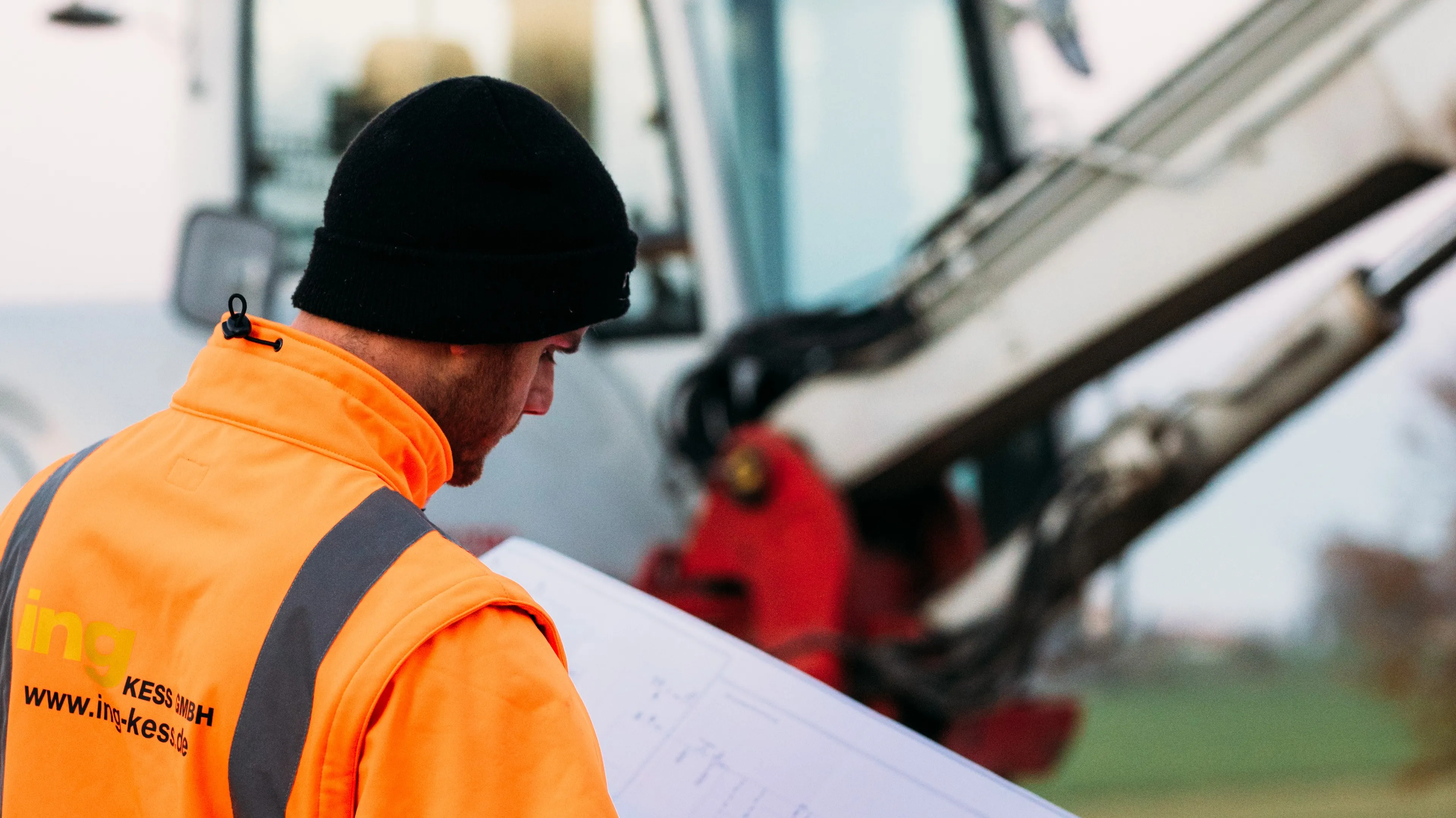Worker in a bright orange vest examines blueprints near construction machinery on a site, wearing a black beanie for warmth.