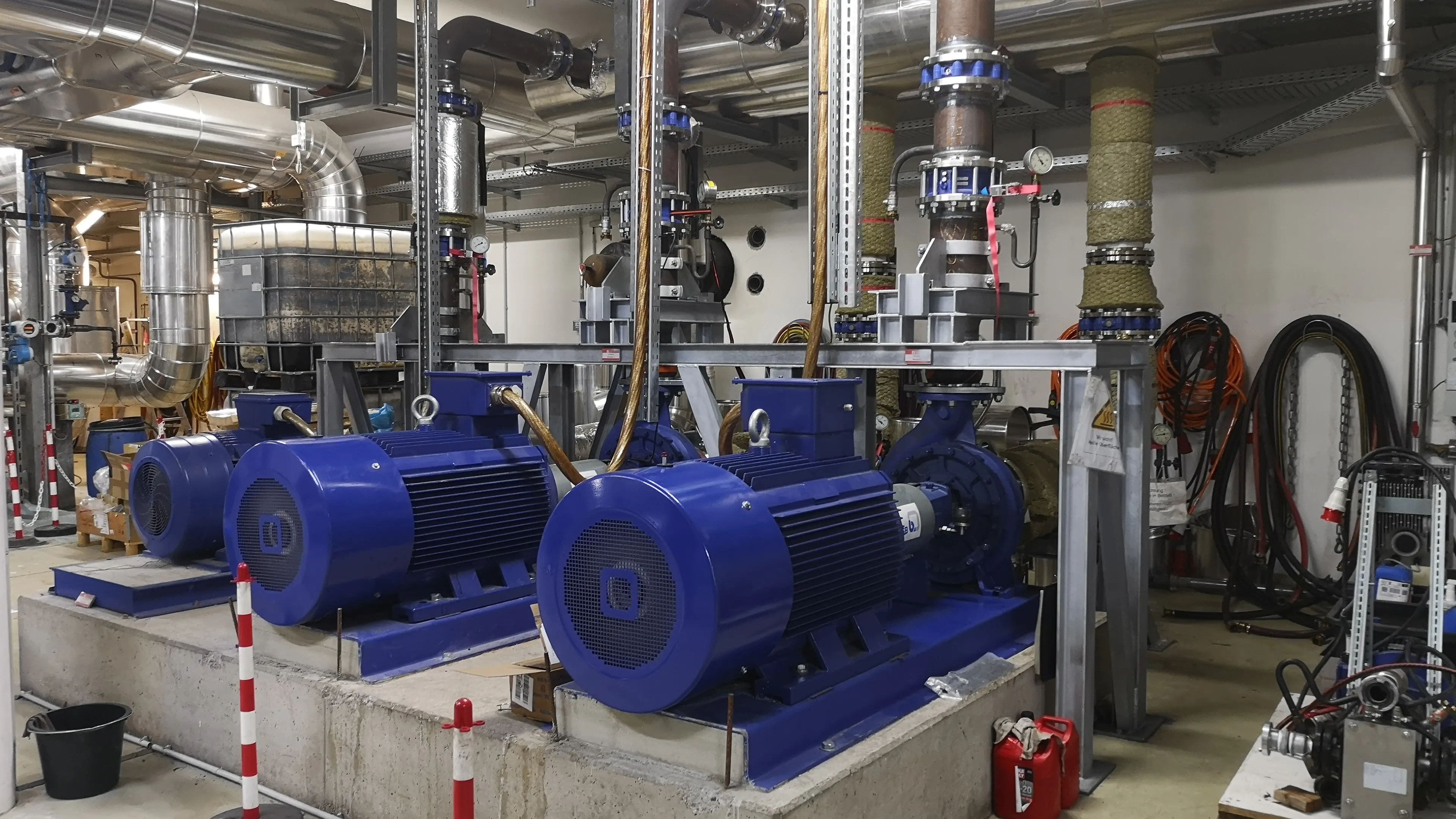 A room containing large blue industrial pumps with pipes overhead, control valves, and safety cones, indicating a mechanical or utility setup.