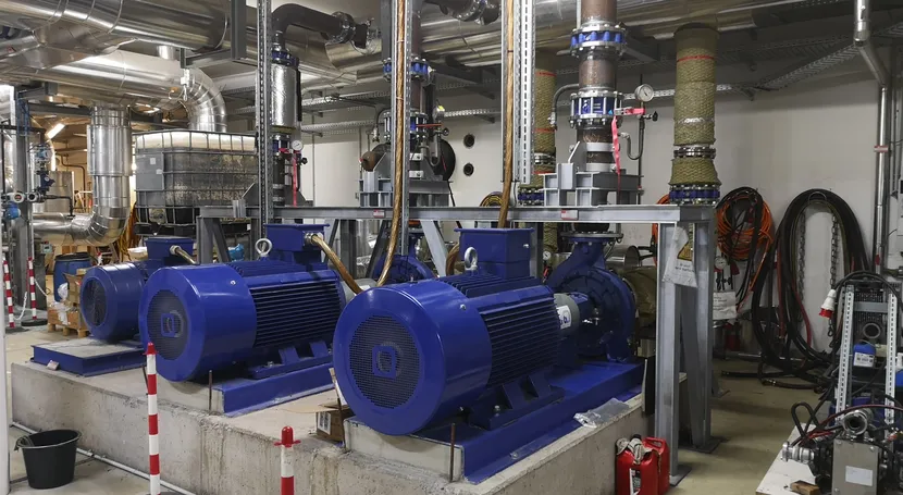 A room containing large blue industrial pumps with pipes overhead, control valves, and safety cones, indicating a mechanical or utility setup.