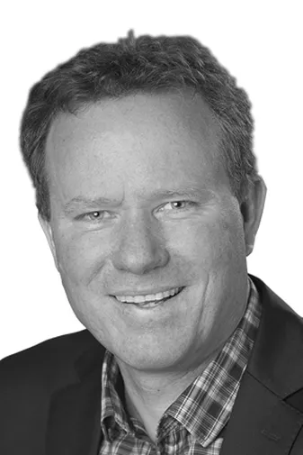 Portrait of a smiling man in a suit jacket and checkered shirt, with short curly hair, against a white background.