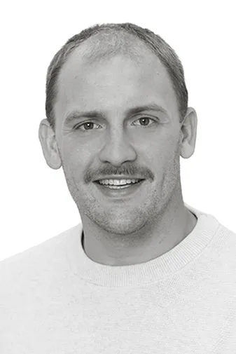 A man with short hair and a mustache smiles, wearing a light-colored sweater, against a plain white background.