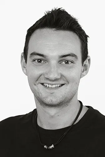 Smiling person with short dark hair, wearing a black shirt and a beaded necklace, against a plain white background.