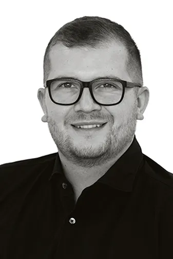 Man with short hair, wearing glasses and a black collared shirt, smiling against a white background.