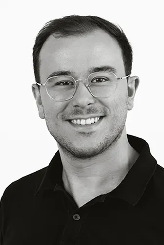 Smiling man with short hair, wearing glasses and a dark collared shirt, set against a plain white background.