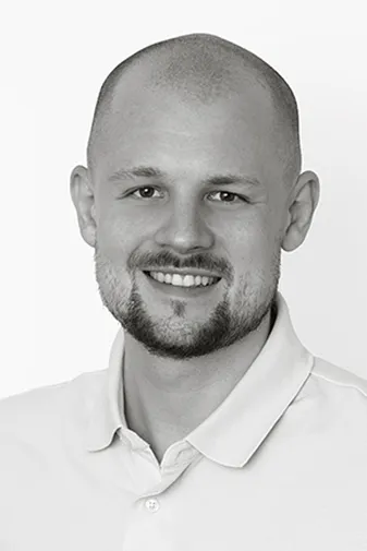 Smiling man with a beard and a shaved head wearing a light-colored polo shirt, set against a plain white background.