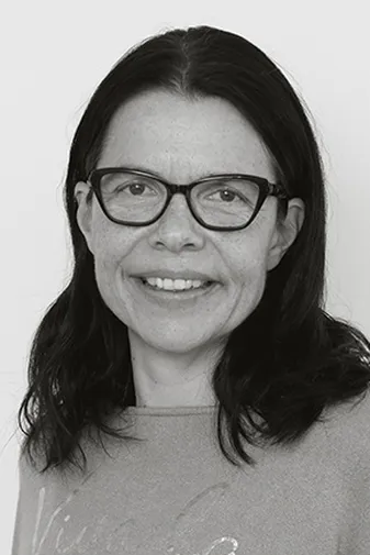 A woman with long dark hair and glasses smiles at the camera. She is wearing a light-colored top, and the background is plain.