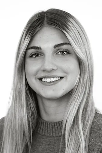 Smiling person with long, straight hair wearing a sweater, set against a plain background. Black and white image.