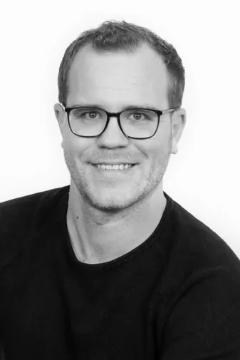 Man with short hair and glasses smiling, wearing a dark sweater, against a plain white background. Black and white photo.