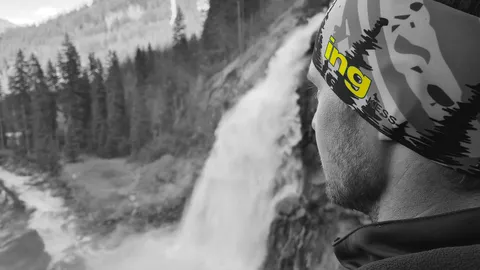 Person in a headband gazes at a waterfall surrounded by a forest, with mountains in the background. Grayscale with selective color on the headband.