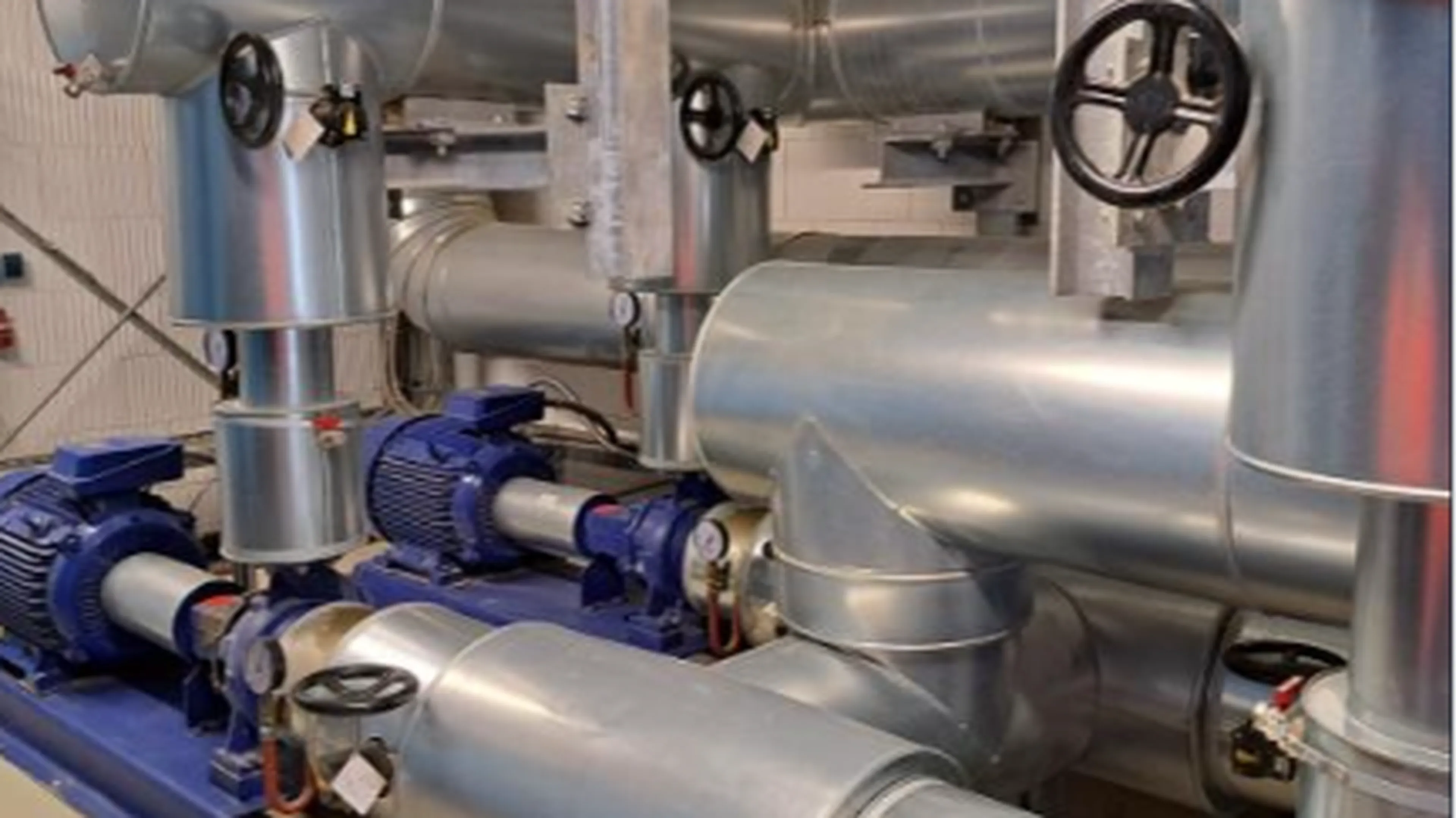Industrial machinery with large metal pipes, blue motors, and black valve wheels in a mechanical room setting.
