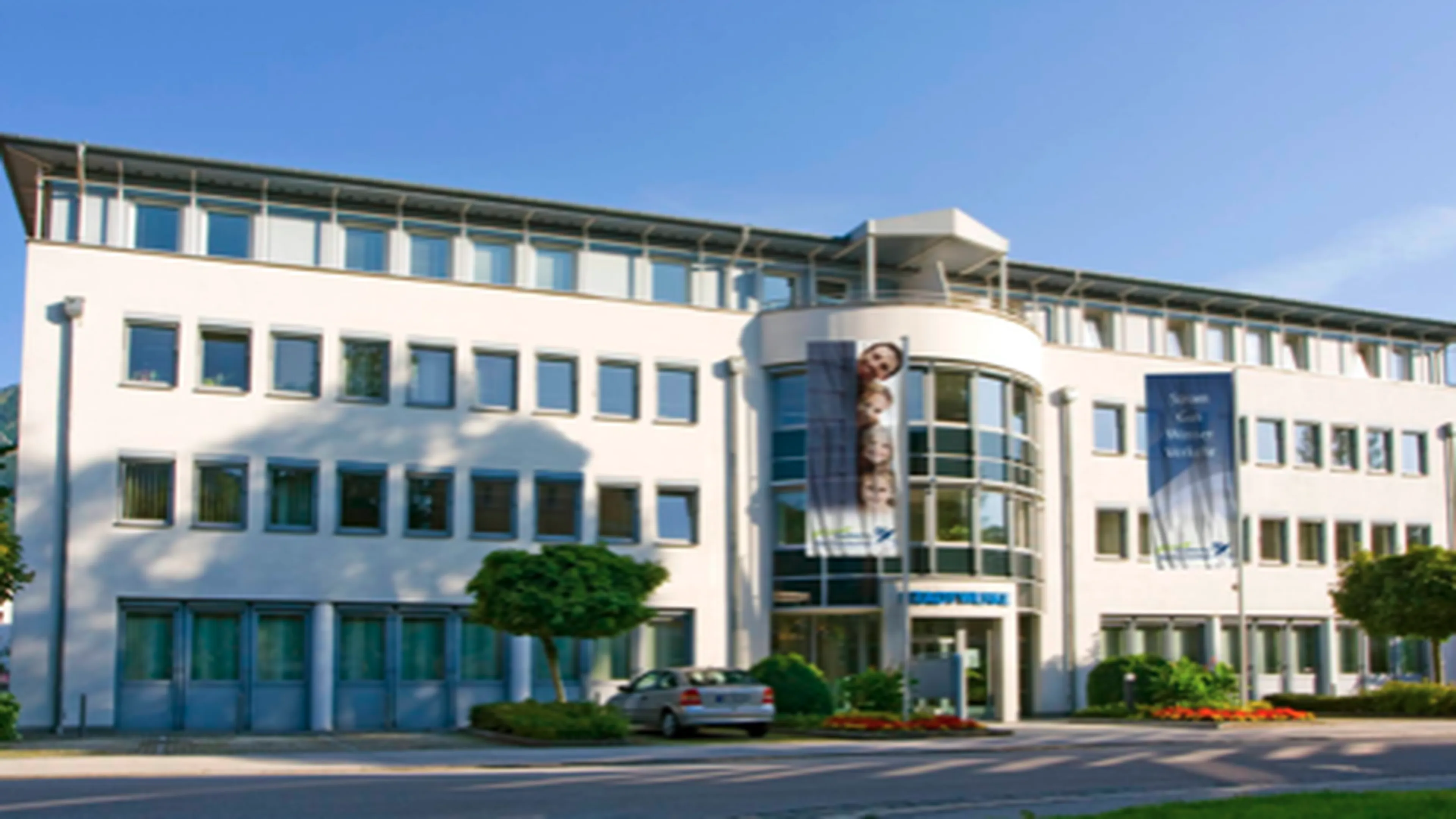 Modern white office building with large windows, banners, and a central glass entrance. Trees and a parked car are visible in front.