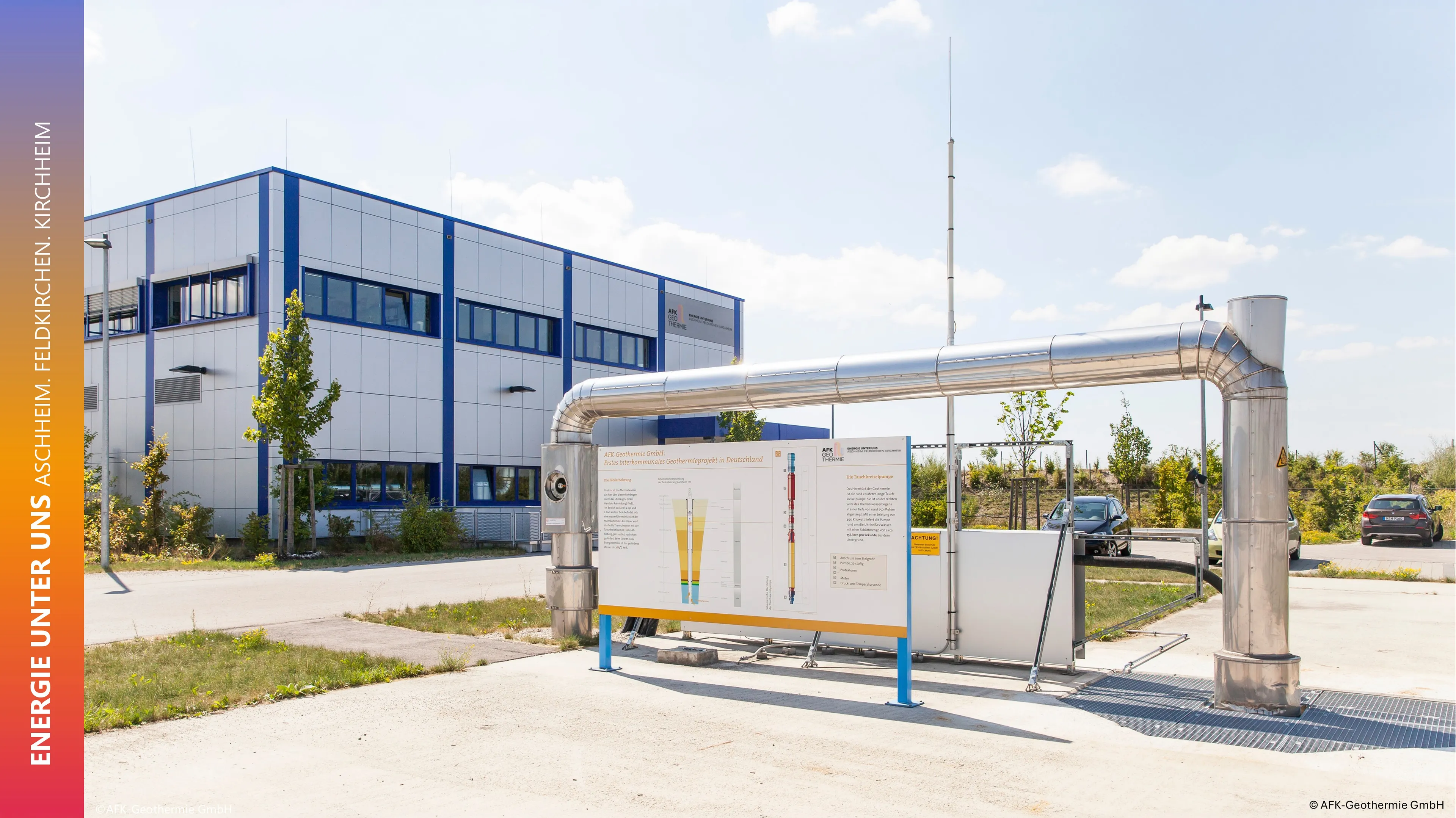 Modern building and geothermal energy display with pipes and diagrams, surrounded by trees and parked cars, under a clear blue sky.