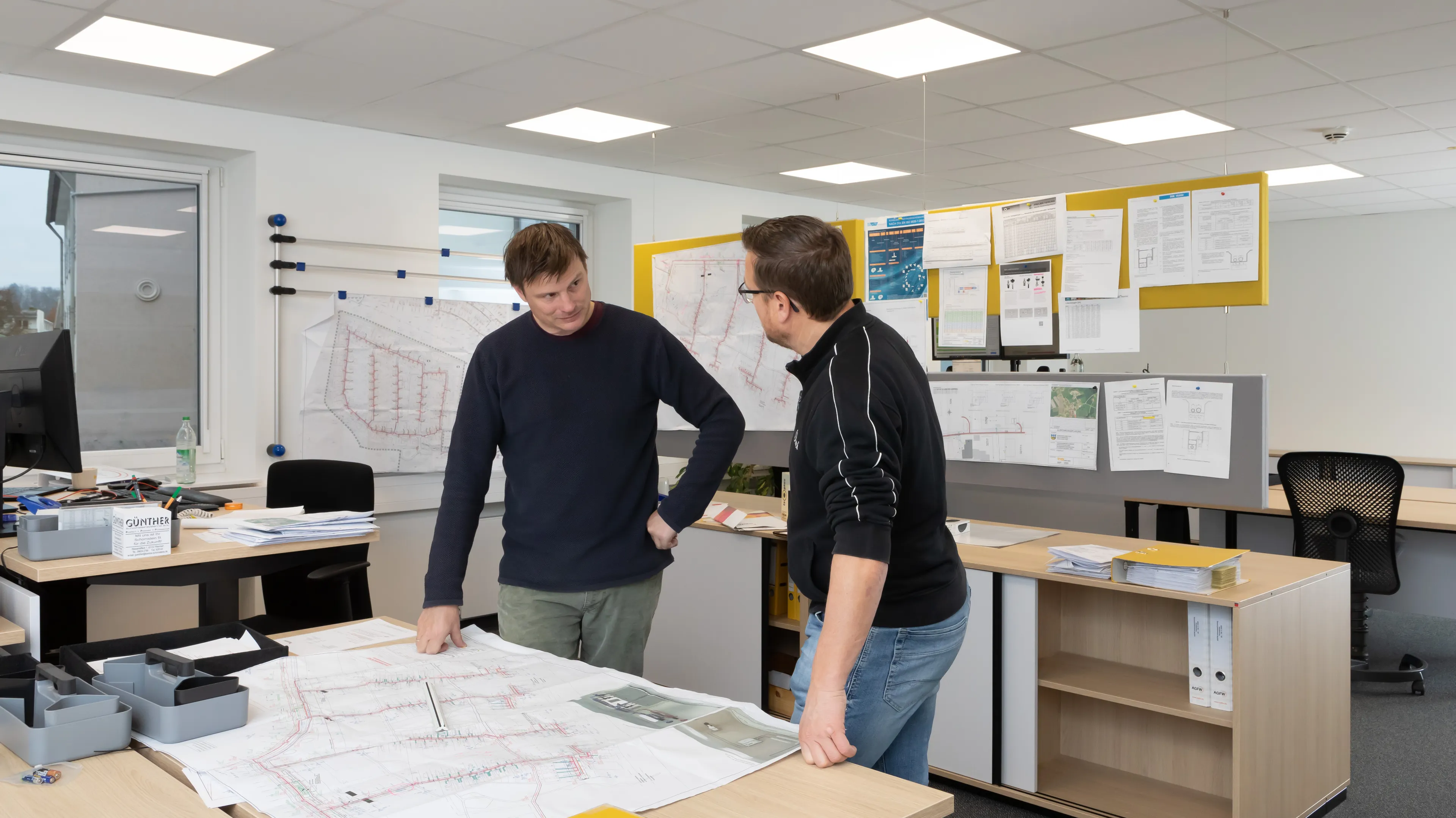 Two men discussing architectural plans in an office with maps and documents on the walls.