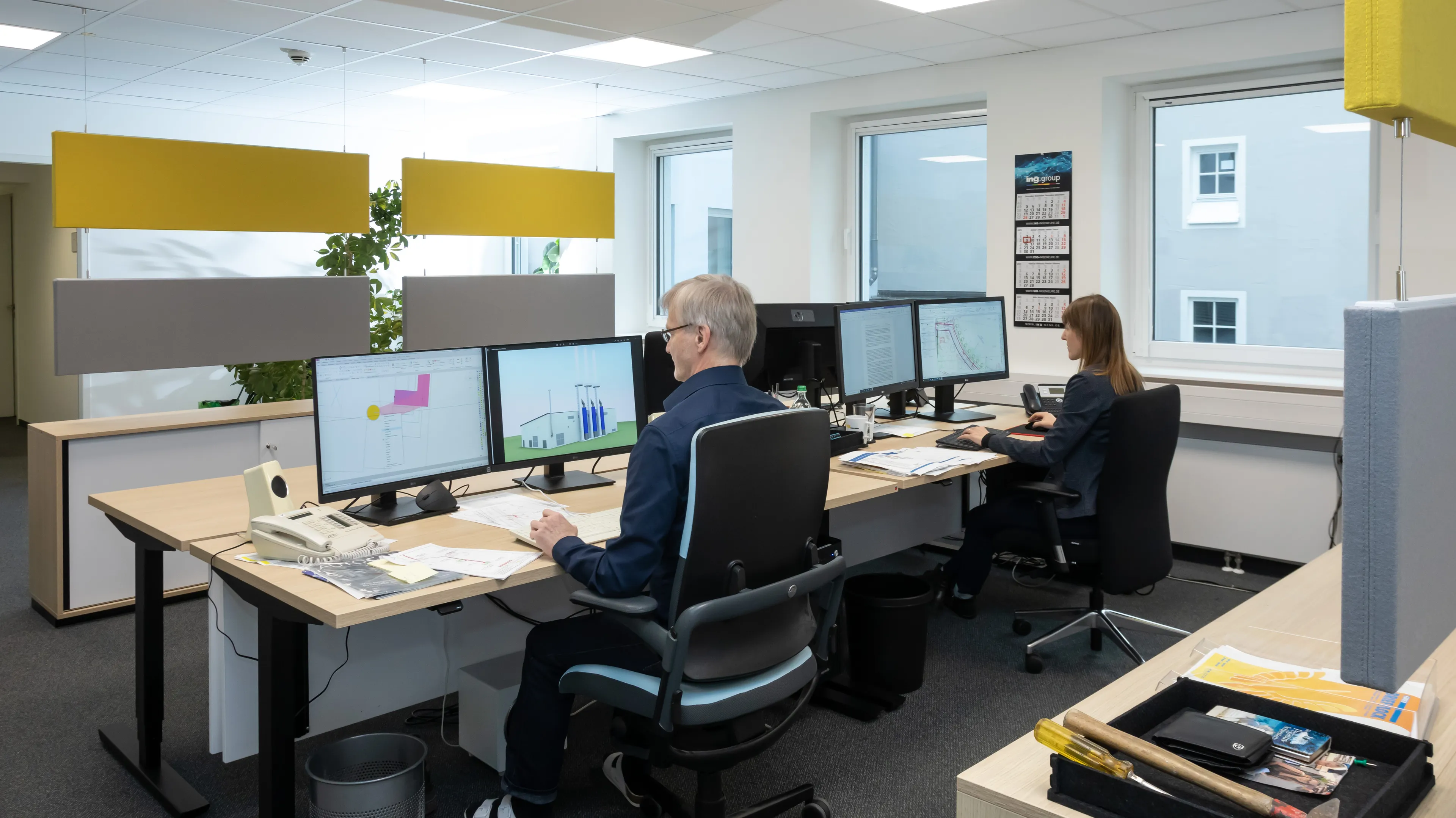 Two people working at desks with multiple computer monitors in a modern office, separated by yellow and gray dividers.