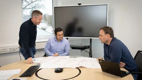 Three men discussing over a map at a round table in a meeting room with a large monitor and a window in the background.