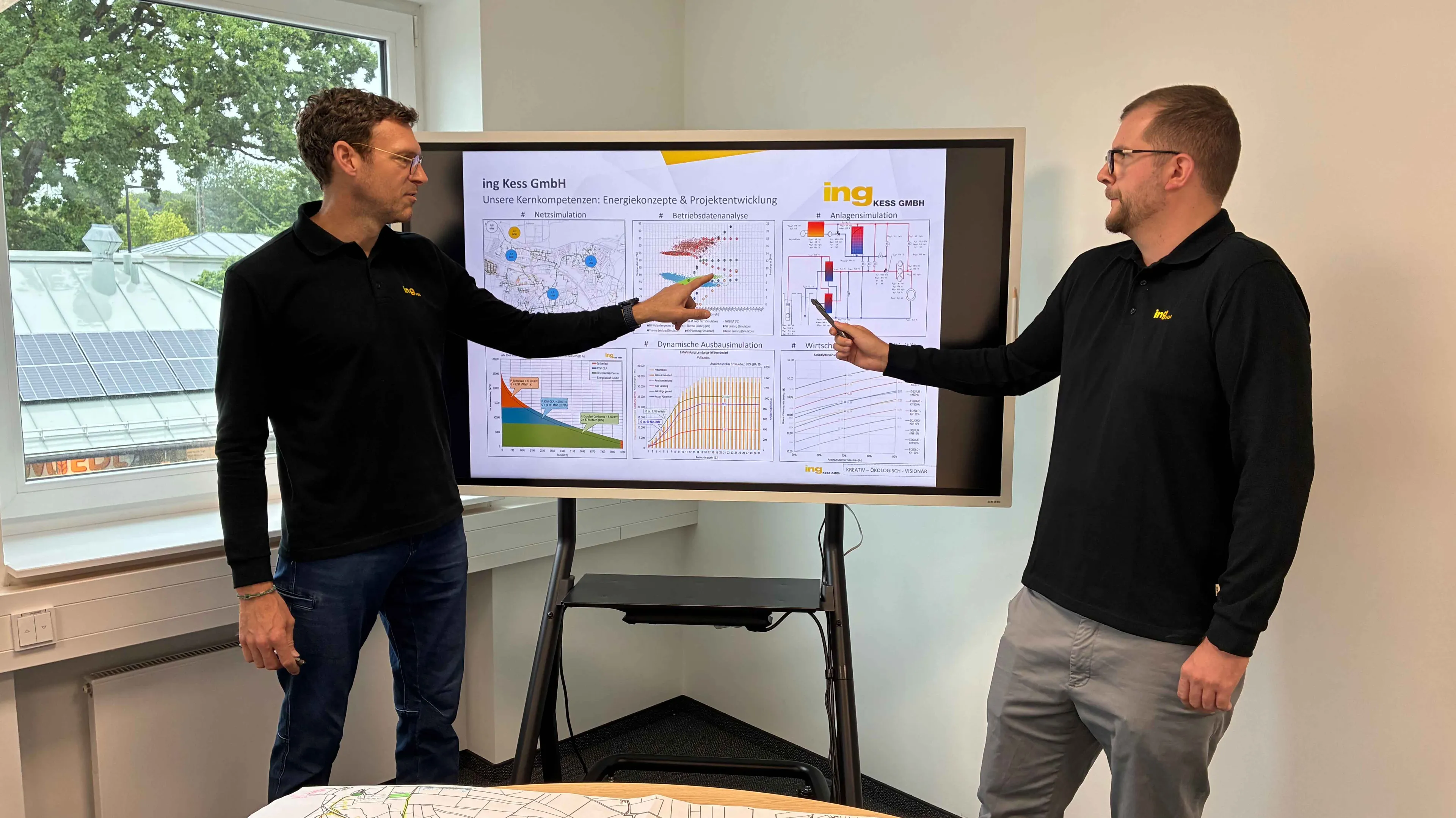 Two men in black shirts discuss charts on a large screen in an office with a window showing solar panels outside.