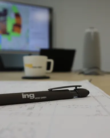 Technische Projektplanung in einem Besprechungsraum: Ein Kugelschreiber liegt auf Planungsunterlagen, während im Hintergrund technische Visualisierungen auf einem Bildschirm dargestellt werden. Das Bild steht für Ingenieurleistungen, Projektmanagement und technische Konzeption.