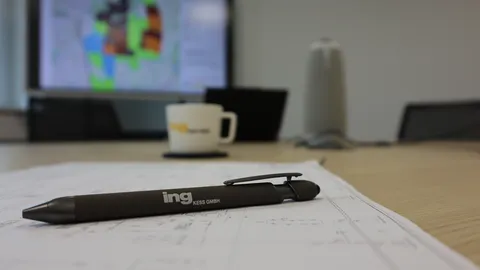 Technische Projektplanung in einem Besprechungsraum: Ein Kugelschreiber liegt auf Planungsunterlagen, während im Hintergrund technische Visualisierungen auf einem Bildschirm dargestellt werden. Das Bild steht für Ingenieurleistungen, Projektmanagement und technische Konzeption.