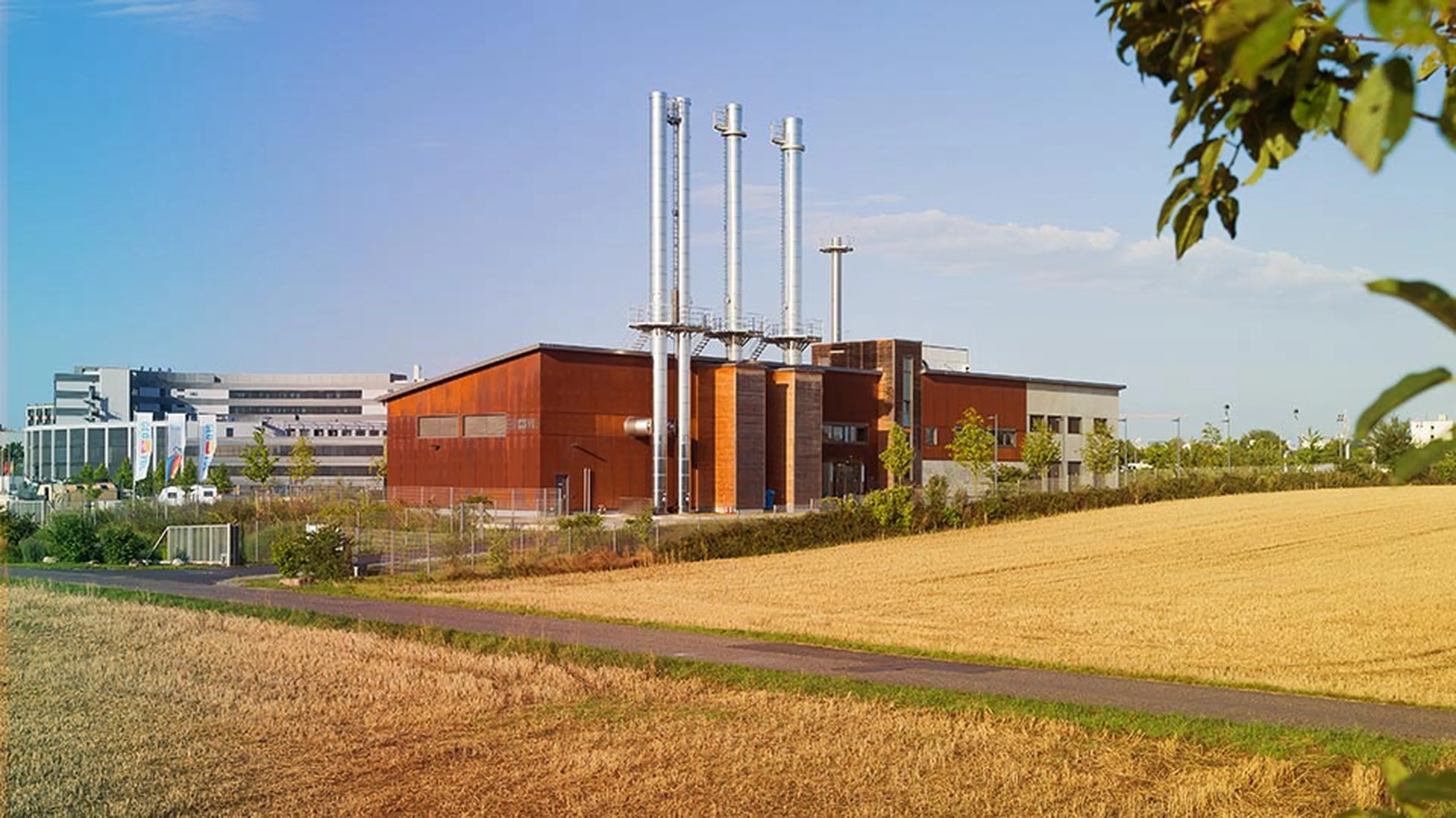 Industrial building with tall chimneys, surrounded by a field and a path, under a blue sky with some clouds.