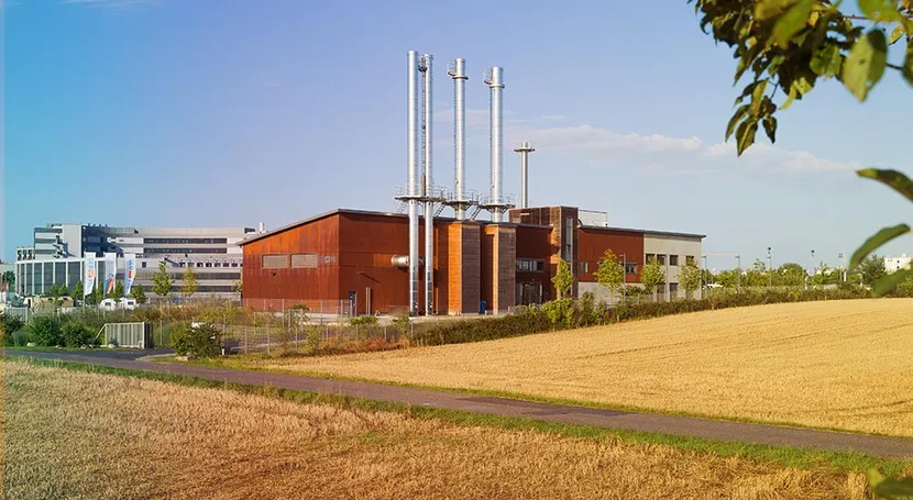 Industrial building with tall chimneys, surrounded by a field and a path, under a blue sky with some clouds.
