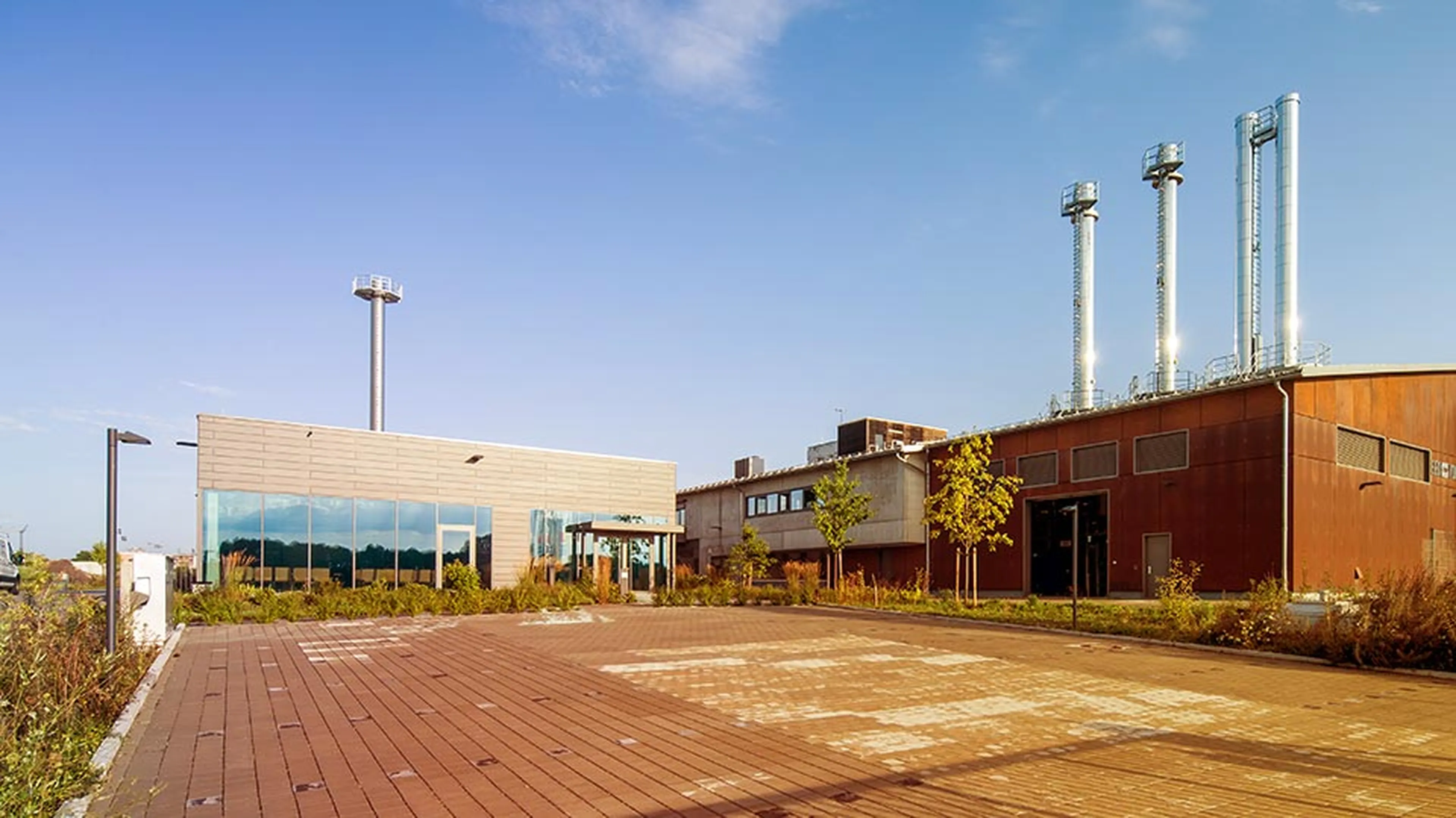 Industrial building with metal chimneys and a modern facade, surrounded by a paved area and sparse vegetation under a clear sky.