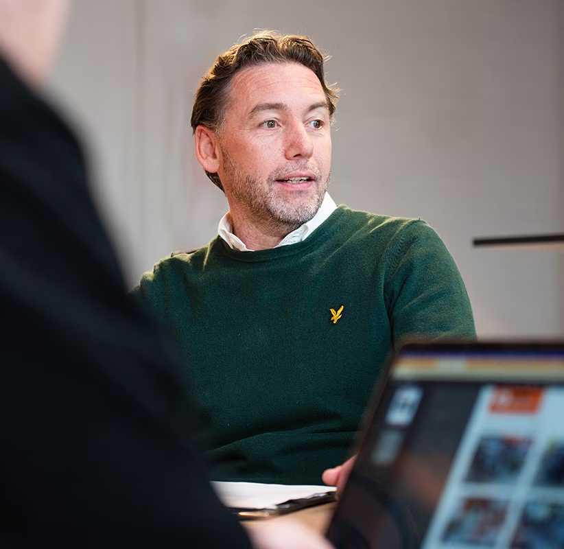 Een man met een baard, gekleed in een groene trui en wit overhemd, zit aan een tafel, kijkt opzij en praat. Een laptop is zichtbaar op de voorgrond, en de schouder van een andere persoon links.