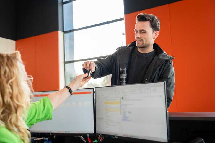 Bij de balie van VWP Shortlease geeft een man in een donkere jas lachend een autosleutel aan een vrouw in een groen shirt. Er staan twee computermonitoren op de balie.