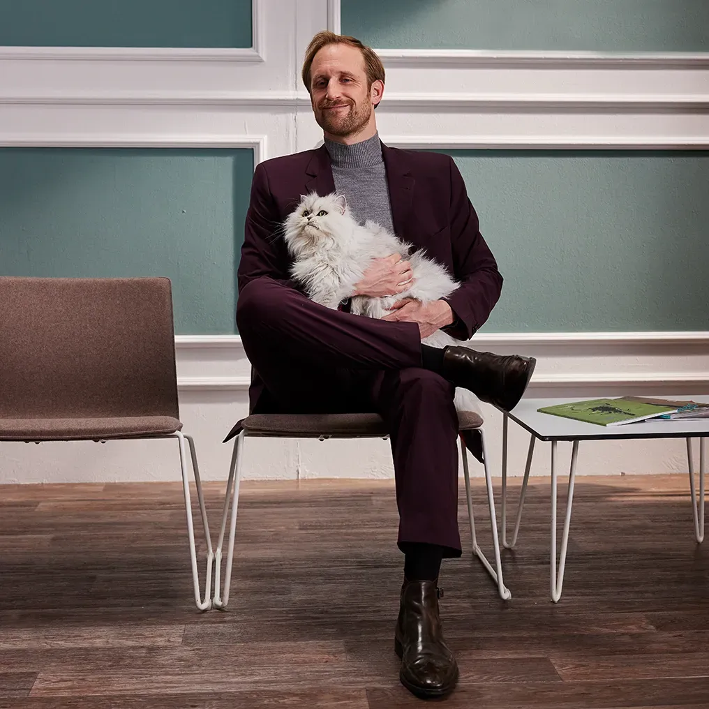Ein Mann in einem lila Anzug sitzt auf einem Stuhl und hält eine flauschige weiße Katze. Er sitzt mit einem entspannten Gesichtsausdruck in einem Raum mit grünen Wänden und Holzboden.