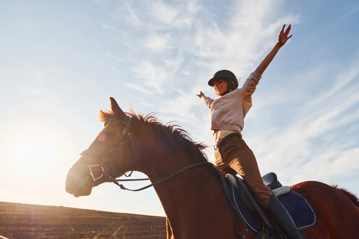 Eine Person reitet freudig auf einem Pferd mit ausgestreckten Armen unter einem strahlenden Himmel, trägt einen Helm und Freizeitkleidung.