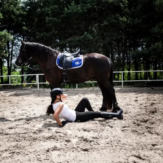 Eine Person in Reitkleidung liegt auf dem Boden neben einem gesattelten Pferd in einer sandigen Arena, mit Bäumen im Hintergrund.