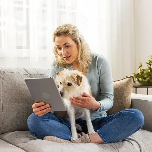 Frau sitzt mit einem Hund auf einer Couch und hält ein Tablet in der Hand. Sie lächelt und trägt einen hellblauen Pullover und Jeans.