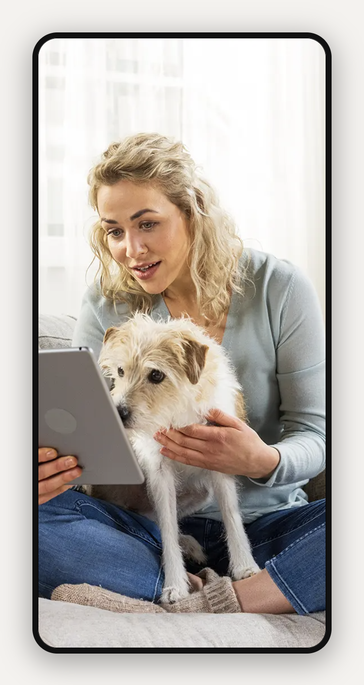 Frau sitzt auf einer Couch, hält ein Tablet und einen kleinen Hund in der Hand. Sie trägt einen hellblauen Pullover und Jeans, mit einem hellen Fenster im Hintergrund.