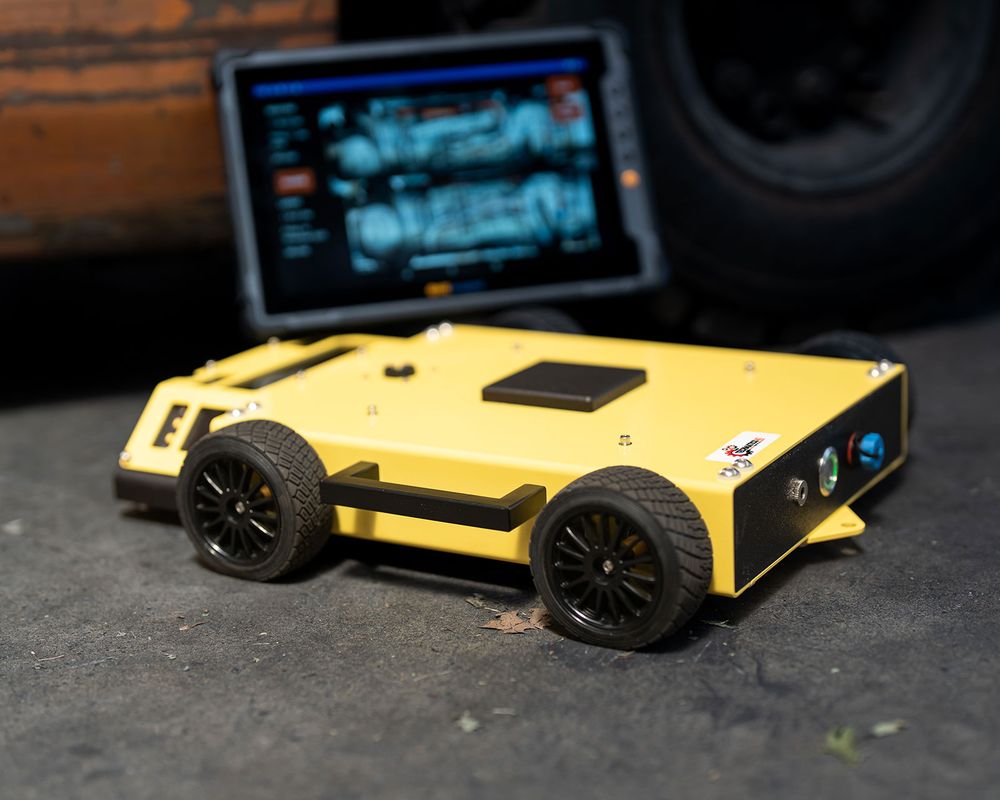 Small yellow four-wheeled inspection robot on concrete floor with a tablet displaying diagnostics behind it.