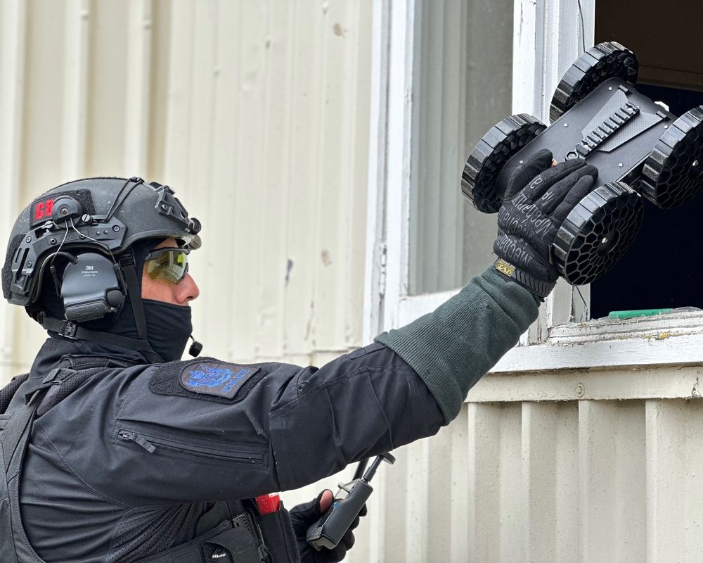 A tactical officer in black gear uses a remote-controlled robot to inspect through a window of a beige building.