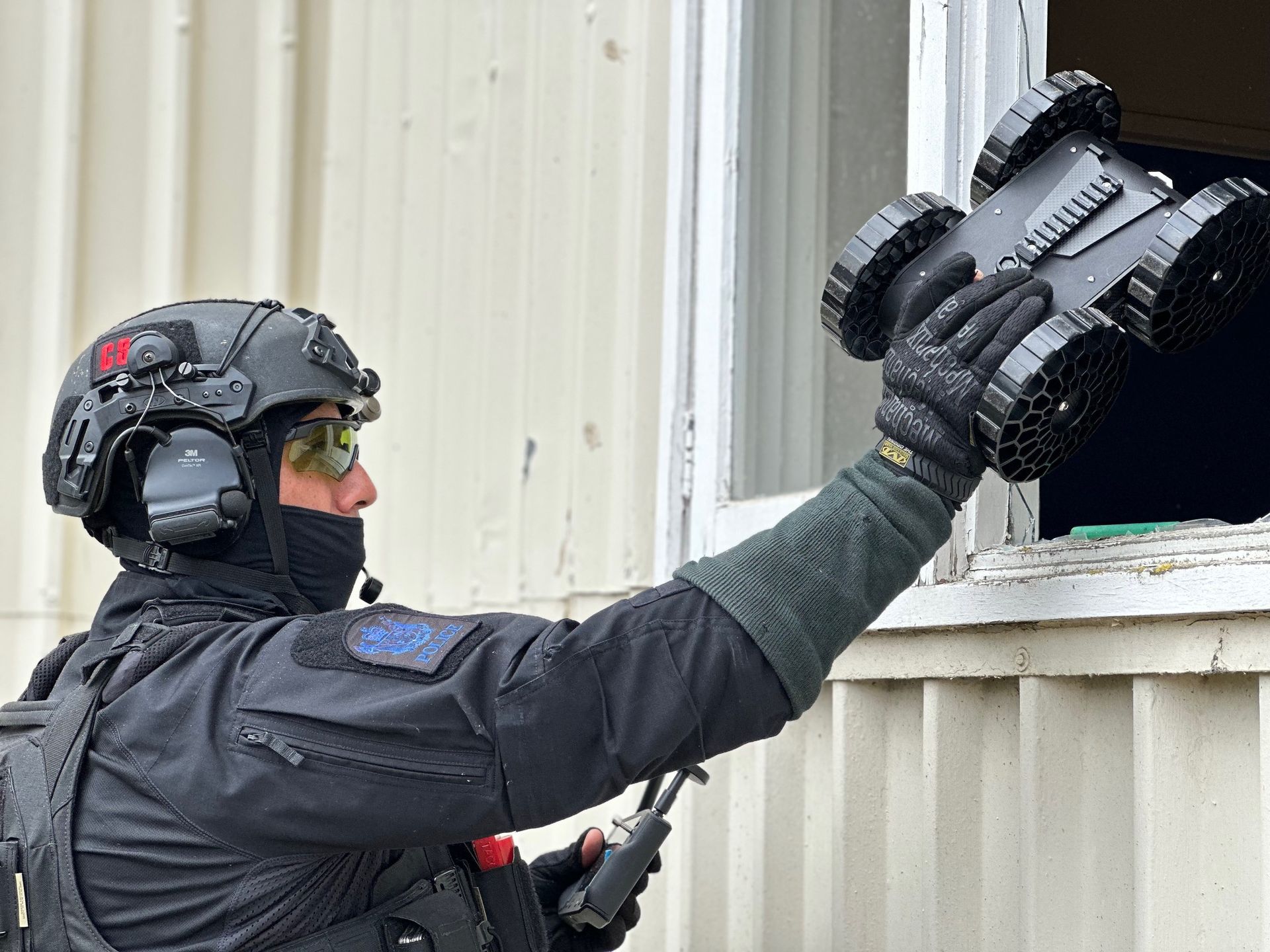 A tactical officer in black gear uses a remote-controlled robot to inspect through a window of a beige building.