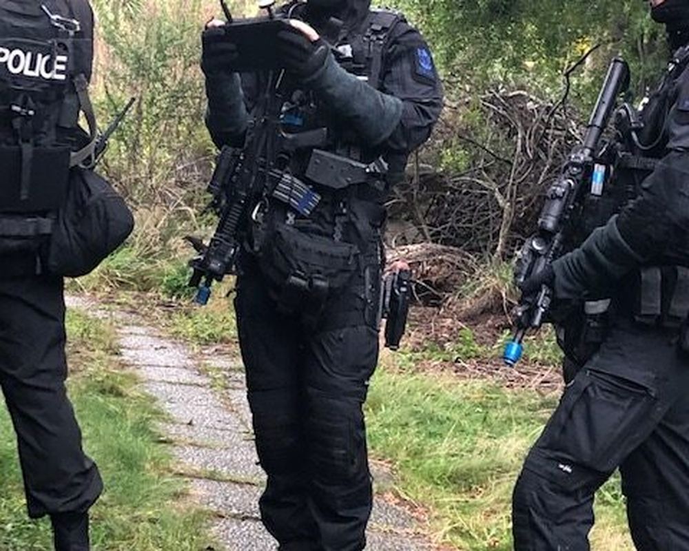 Three police officers in tactical gear stand on a stone path in a wooded area. One officer is writing on a notepad.