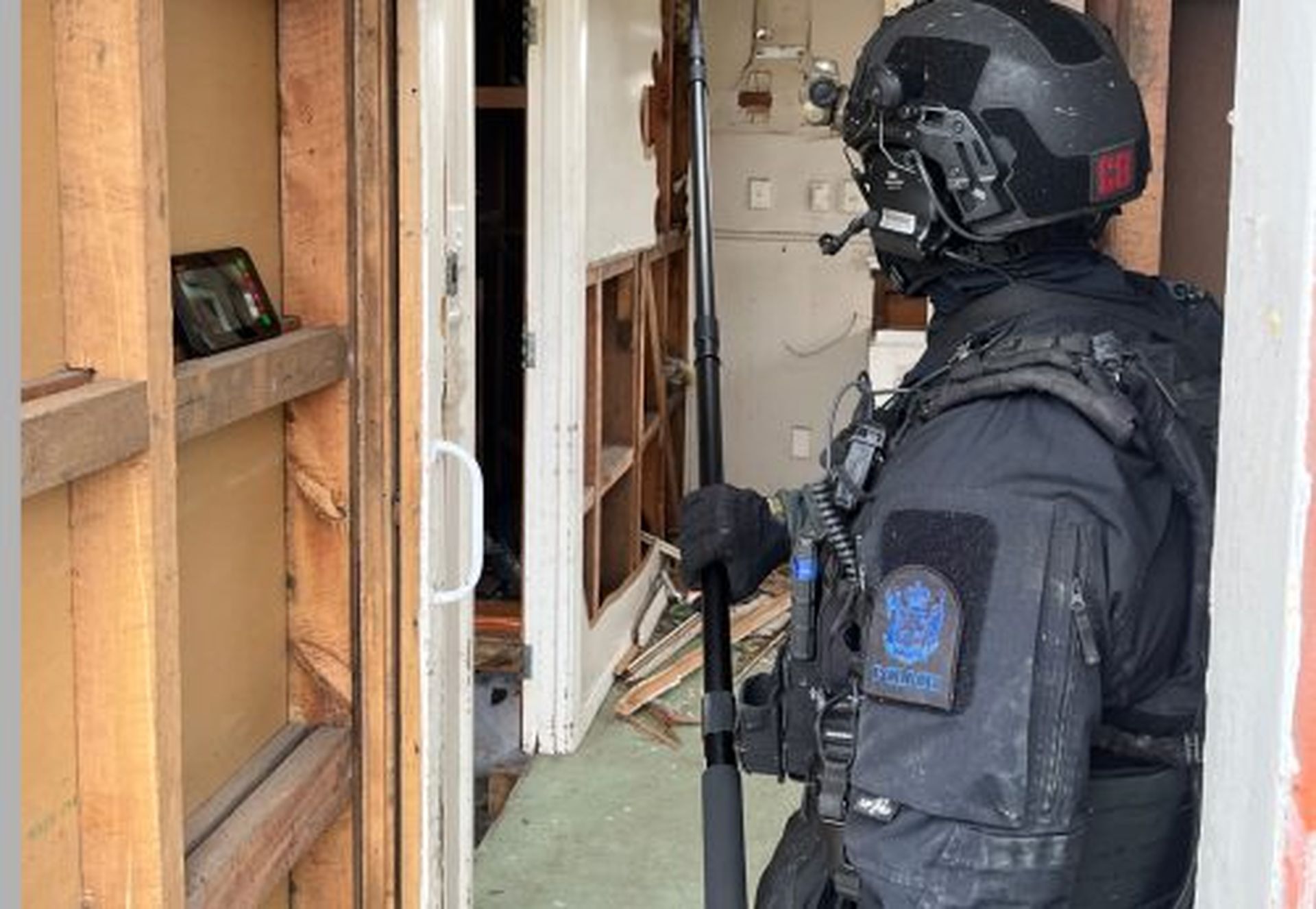 A police officer in tactical gear holds a battering ram, standing at a partially open door of a cluttered room.