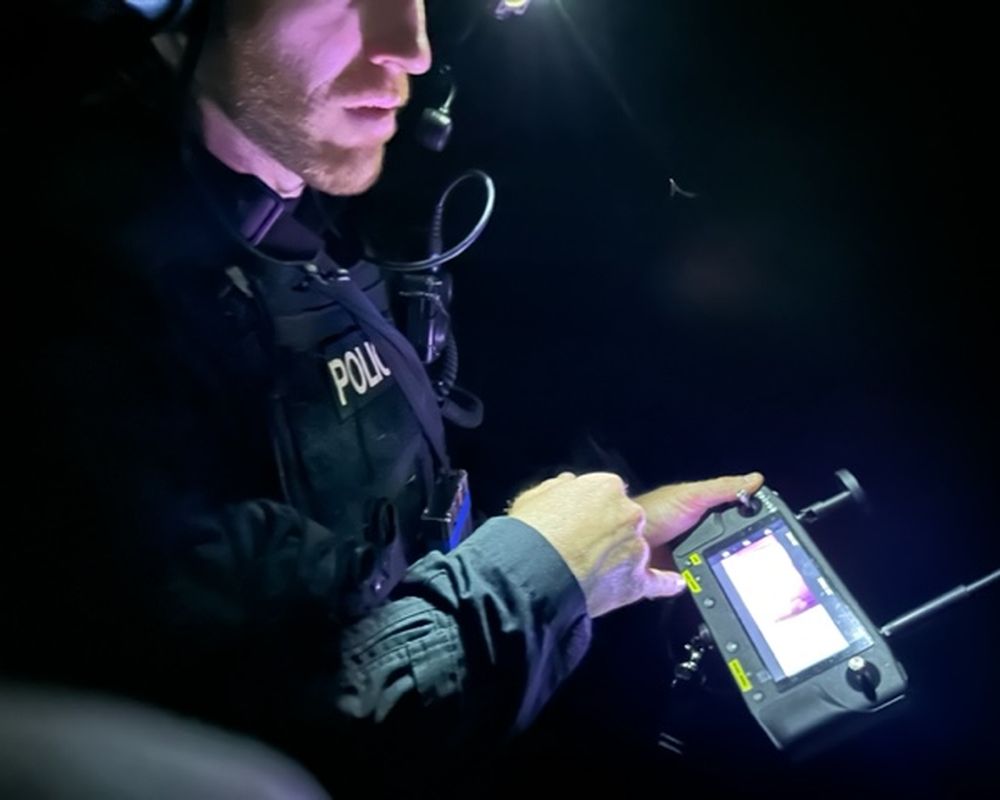 A person in police gear uses a handheld device with a screen, illuminated by a headlamp, in a dark setting.