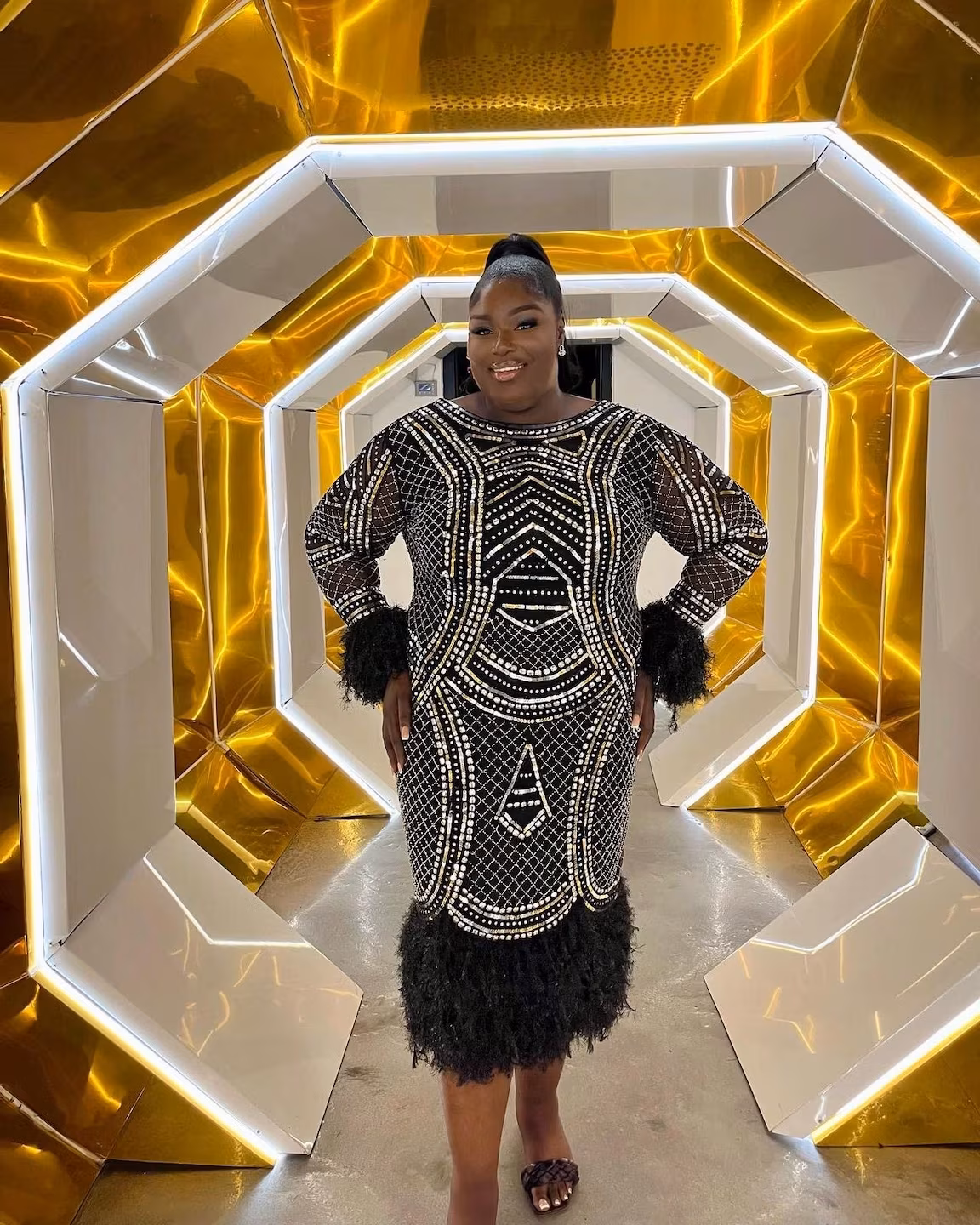 agt-Mitarbeiterin in einem schwarzen, perlenbesetzten Kleid mit Fellmanschetten steht lächelnd in einem futuristischen, golden beleuchteten Tunnel.