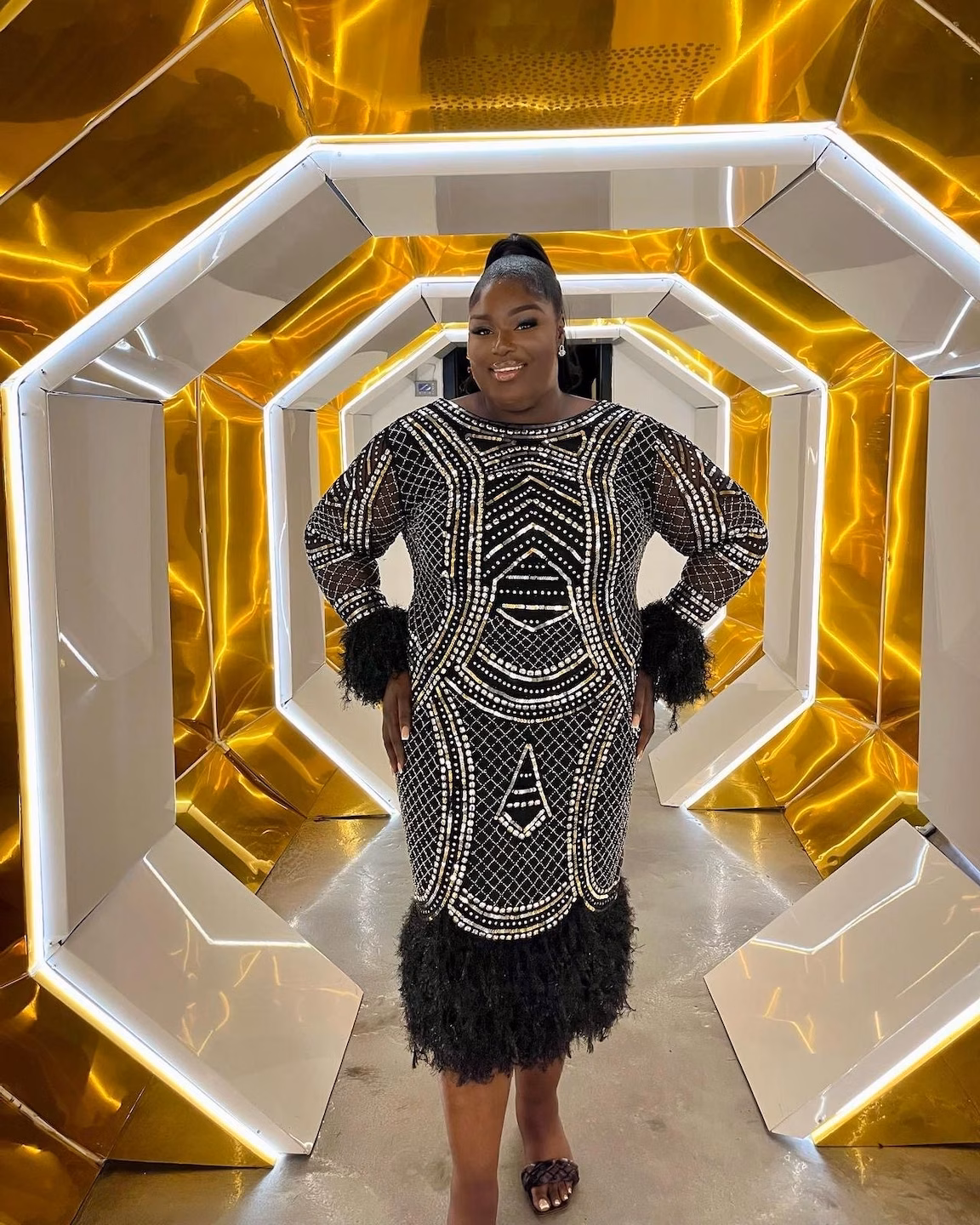 agt-Mitarbeiterin in einem schwarzen, perlenbesetzten Kleid mit Fellmanschetten steht lächelnd in einem futuristischen, golden beleuchteten Tunnel.