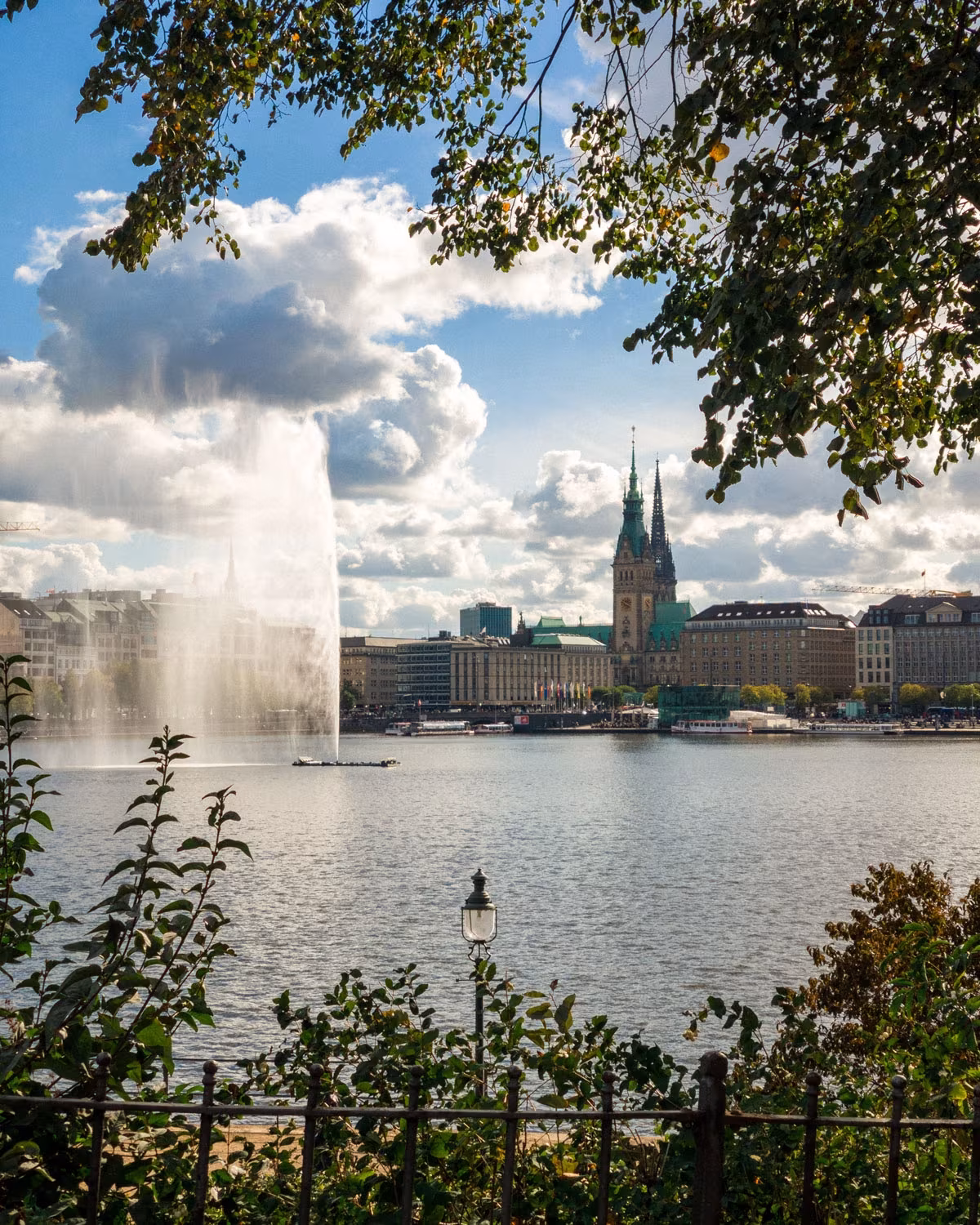 Blick über die Hamburger Binnenalster mit der Alsterfontäne, umrahmt von Bäumen, und dem Rathaus im Hintergrund unter einem wolkenreichen Himmel.