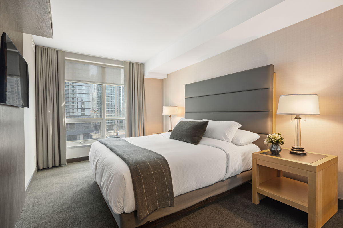 Das letzte Bild zeigt ein helles, modern eingerichtetes Hotelzimmer mit einem großen Doppelbett, zwei Nachttischen samt Lampen und einem großen Fenster im Hintergrund, durch das man auf ein städtisches Gebäude blickt.