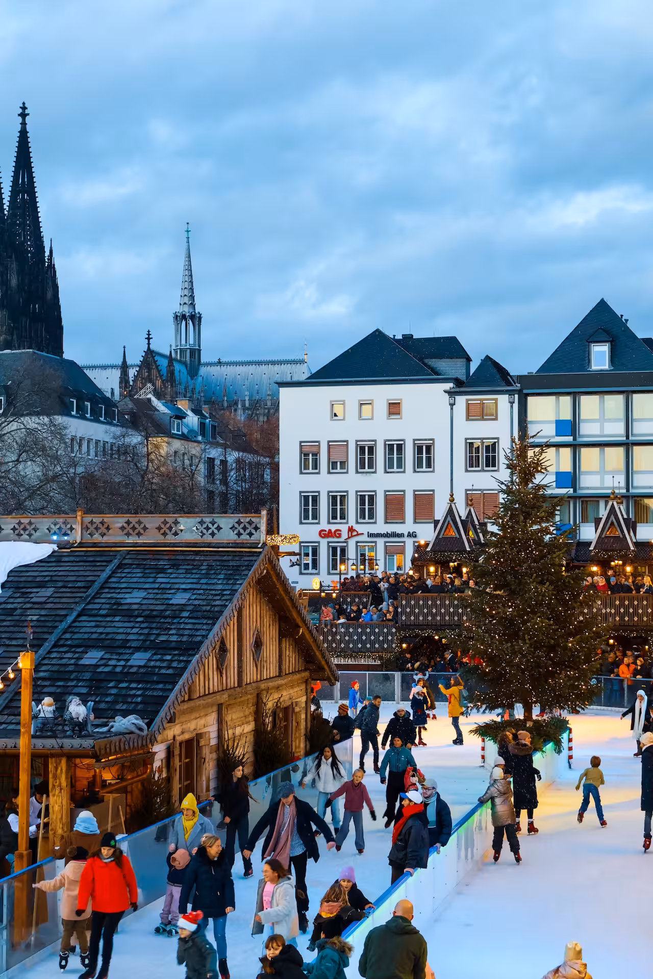 Eislaufbahn auf einem Kölner Weihnachtsmarkt mit beleuchtetem Tannenbaum und Holzhütten, im Hintergrund der Kölner Dom.
