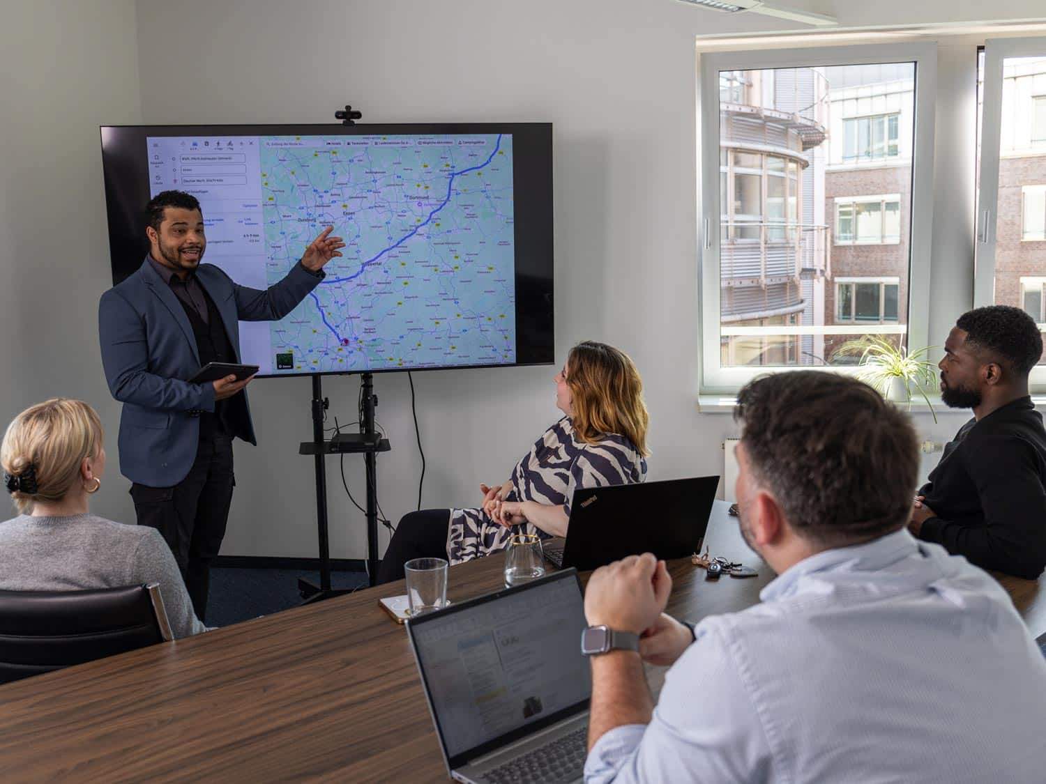 Teammeeting im Besprechungsraum: Ein Mitarbeiter präsentiert eine Route auf einem Bildschirm, während Kolleginnen und Kollegen die Planung verfolgen.