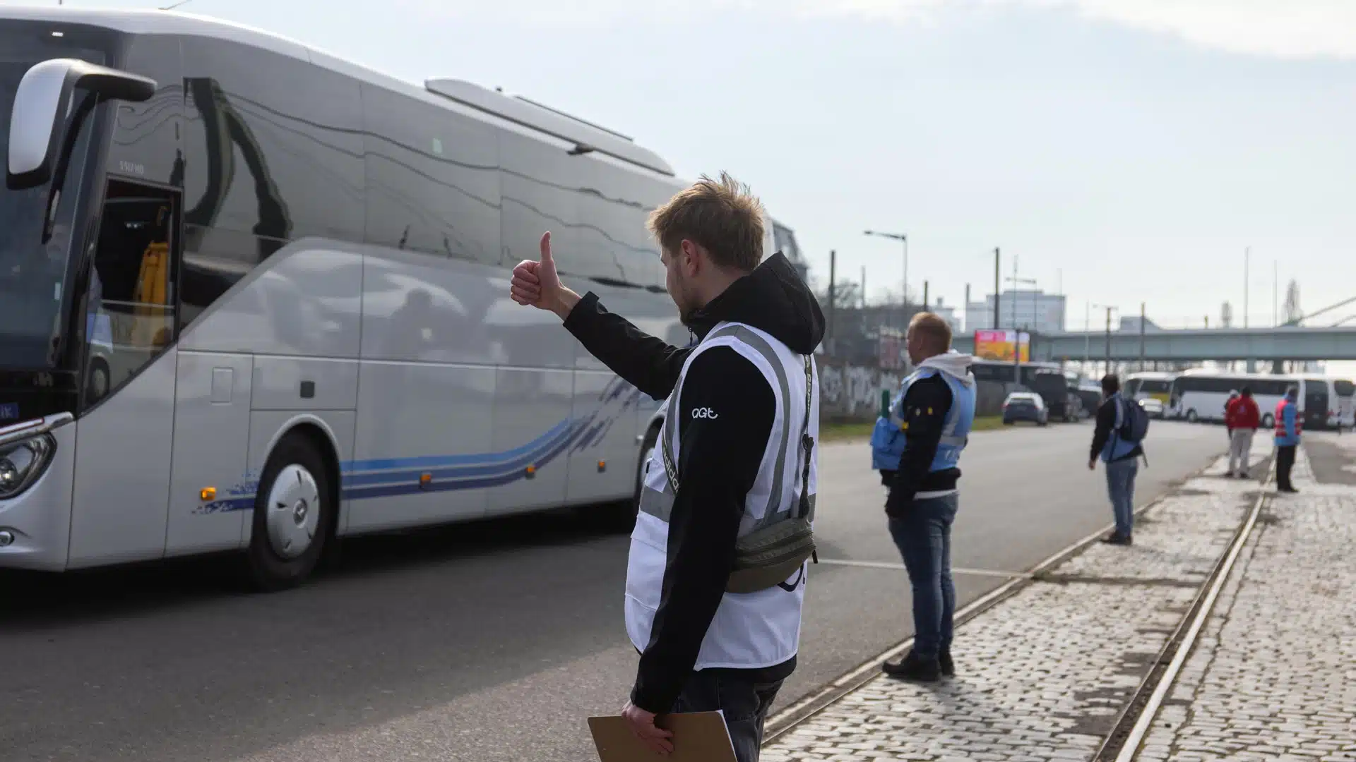 Ein Mann in einer weißen Weste mit agt-Logo gibt einem vorbeifahrenden Reisebus das Daumen-hoch-Zeichen, während im Hintergrund weitere Mitarbeiter an einer Haltestelle stehen.