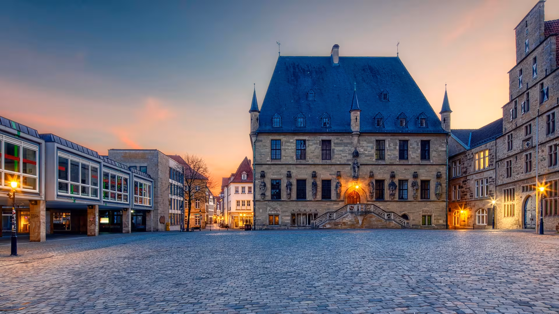 Historisches Rathaus von Osnabrück mit Freitreppe und steilem Schieferdach am leeren, gepflasterten Marktplatz in der Abenddämmerung.